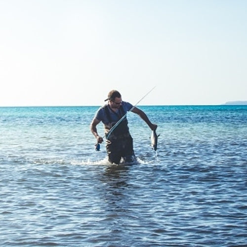zu sehen ist ein Fliegenfischer der im Meer steht und einen Fisch in der Hand hält.