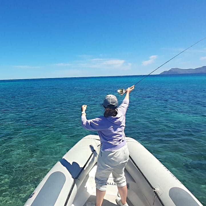 zu sehen ist eine Frau von hinten, stehend in einem Schlauchboot auf dem Meer, die eine Fliegenrute wirft
